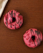 Zwei pink glasierte Donuts mit dunkler Schokoladenlinie und roten Krümeln liegen diagonal auf einer dunklen Holzoberfläche; links oben ein helles Tuch im Anschnitt.
