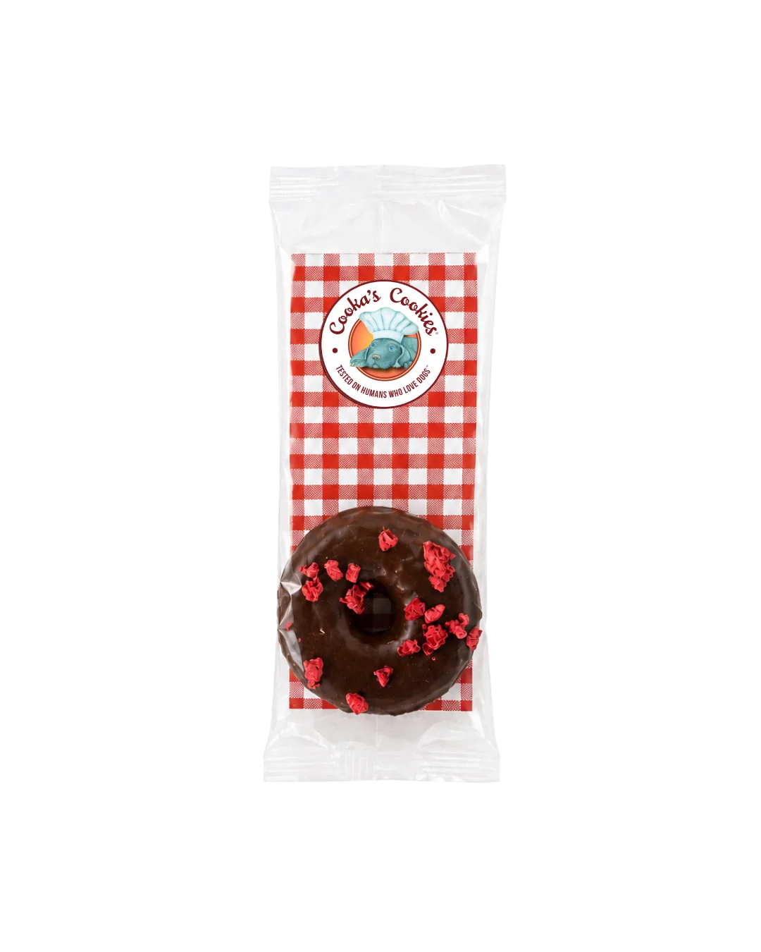 Donutförmiges Hundeleckerli in transparenter Verpackung, dunkelbraun glasiert mit roten Krümeln und dunkler Schokoladenlinie; oben rundes „Cooka’s Cookies“-Etikett auf rot‑weiß kariertem Hintergrund.