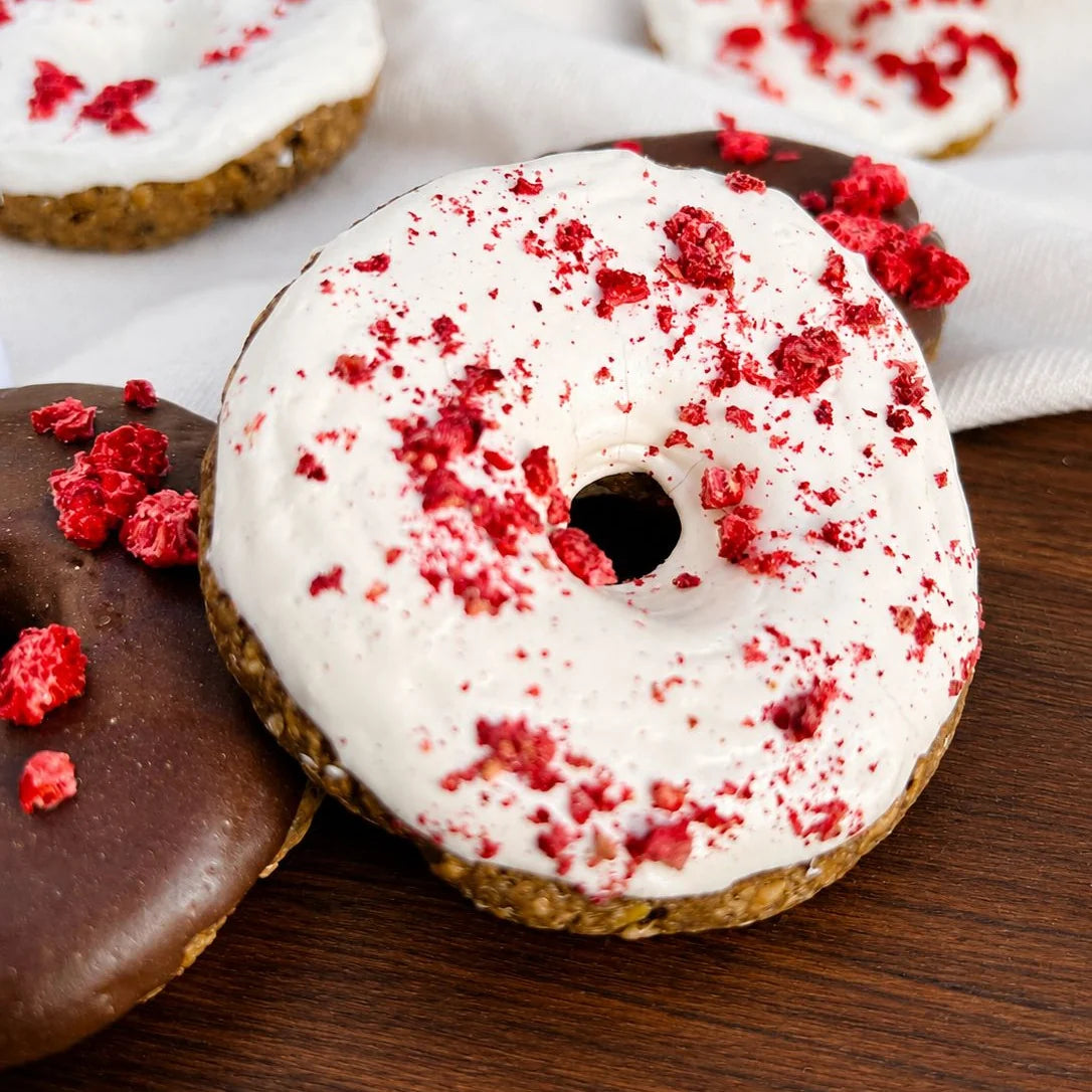 Zwei Donuts auf dunklem Holz, einer weiß glasiert mit roten Stückchen, einer schokoliert mit roten Sprenkeln