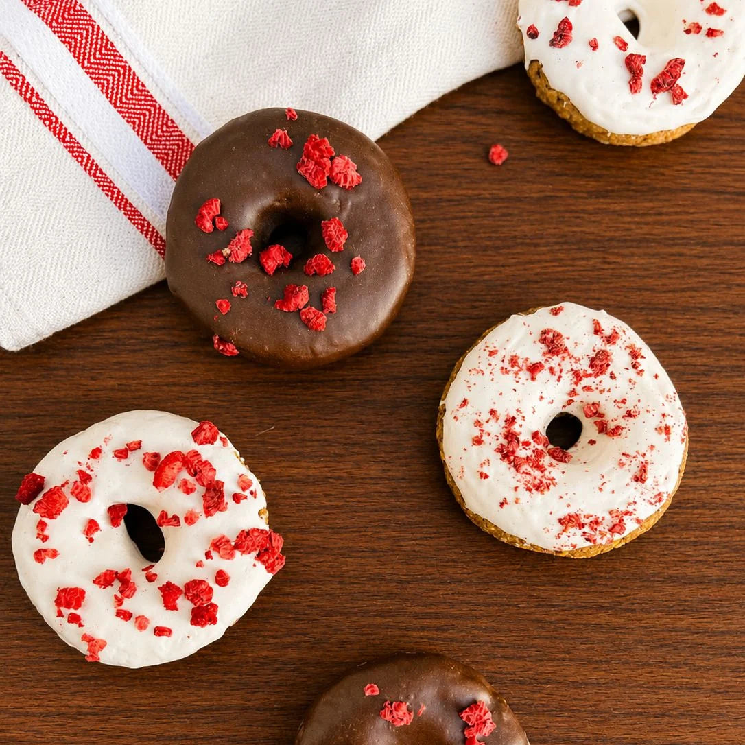 Fünf kleine Donuts auf dunklem Holz, drei weiß glasiert mit roten Stückchen, zwei schokoliert mit roten Sprenkeln; daneben ein weißes Tuch mit rotem Streifen.