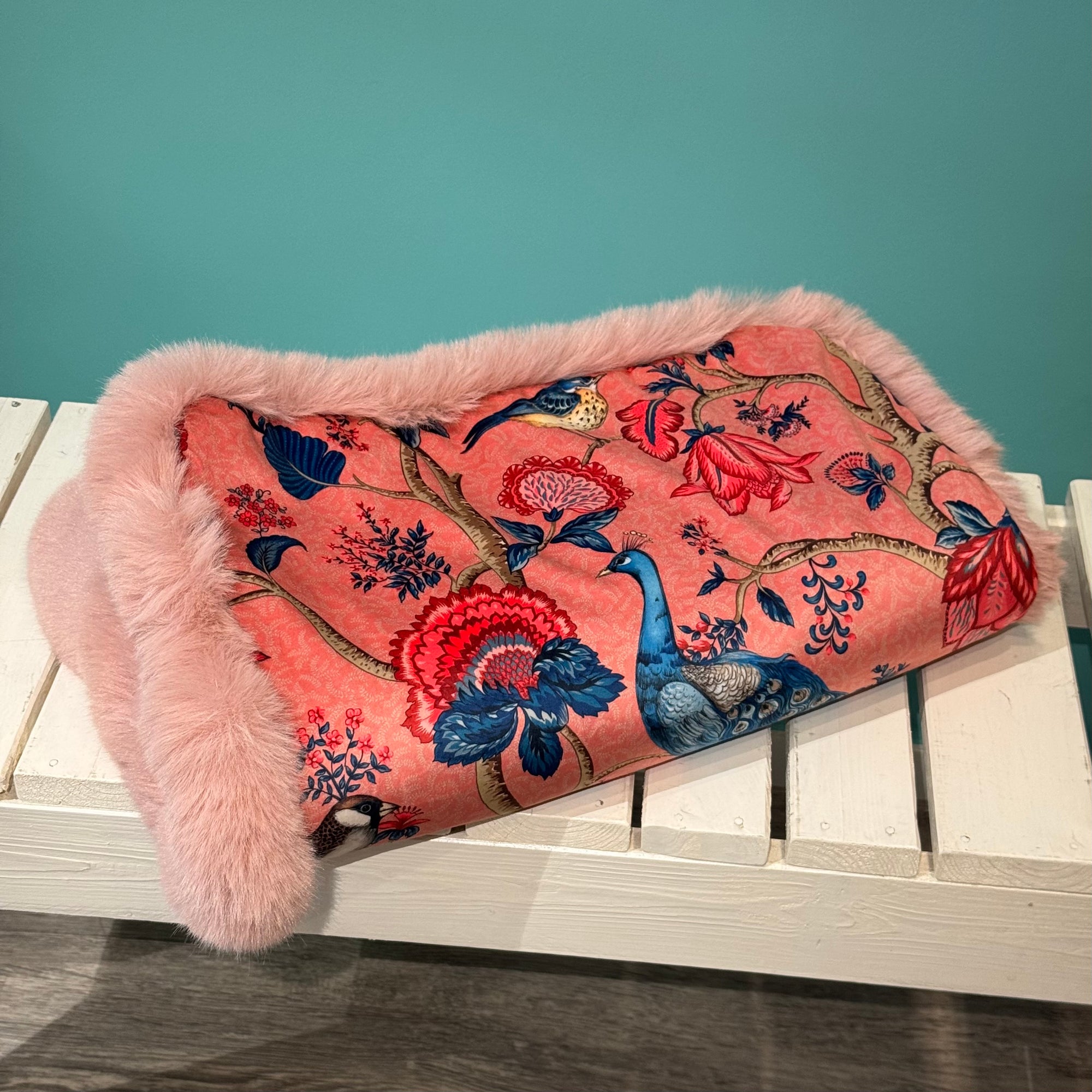Eine Hundeecke mit Kunstfell in Rosa und Samtstoff Pfau  auf einer weißen Bank 