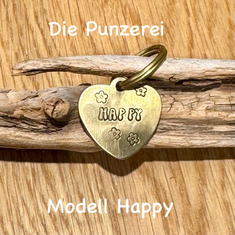 Handgepunzte Hundemarke aus Messing mit Namen „Happy“ und Blumenmotiv, herzförmiges Design an Schlüsselring befestigt, präsentiert auf Holzuntergrund