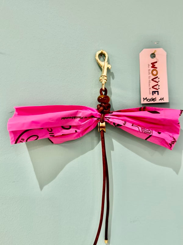 Handgefertigter Isarhund Kotbeutelhalter aus Leder mit goldfarbenem Karabinerhaken, befestigt an einer Wand, mit pinken Kotbeuteln und Markenanhänger, Made in Bavaria