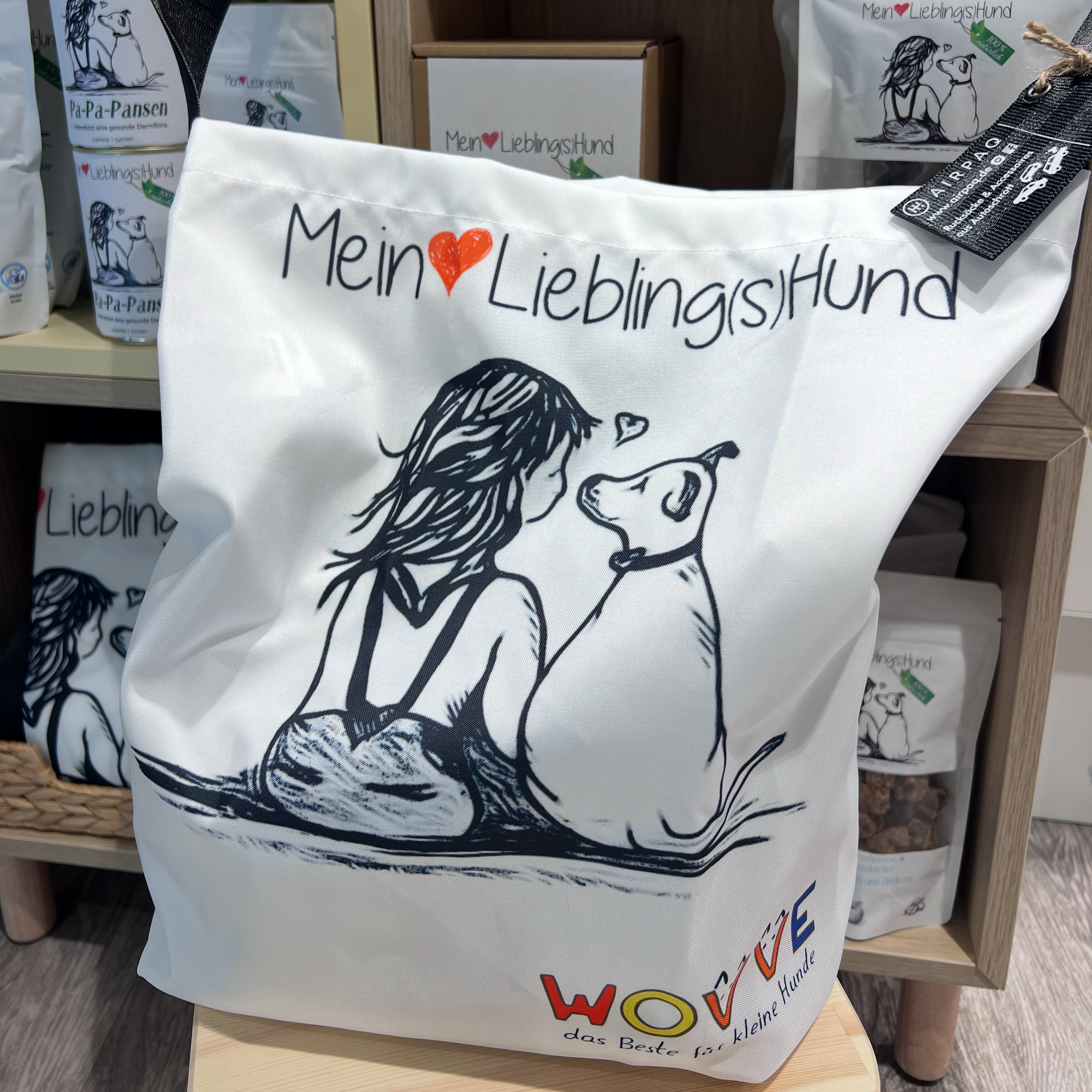 Starterbüdel „Mein Lieblingshund“ mit Hundefutter und Snacks in weißer Tasche aus recyceltem Airpaq-Material, illustriert mit Hund-und-Mensch-Motiv, präsentiert im Regal