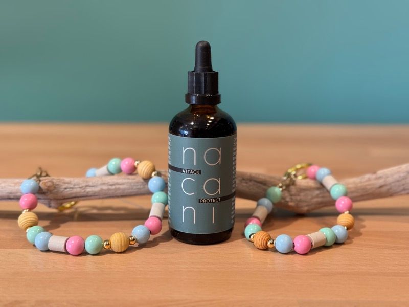 „Nacani Attack Protection Öl mit Pipette, dekoriert mit zwei bunten Anti-Zecken Armbändern aus Holzperlen auf Holzast, Produktfoto“